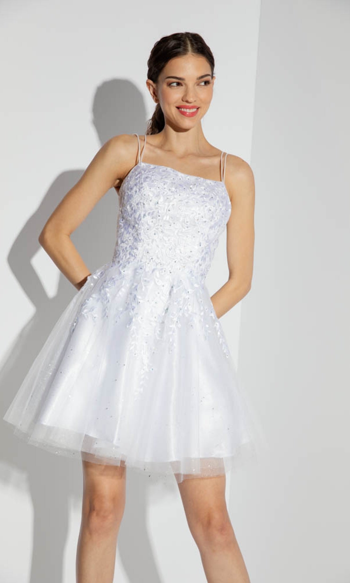 A woman wears the Eureka 9727 Lace-Up Short Prom Dress—white, a-line with thin straps and floral details—against a plain light background.